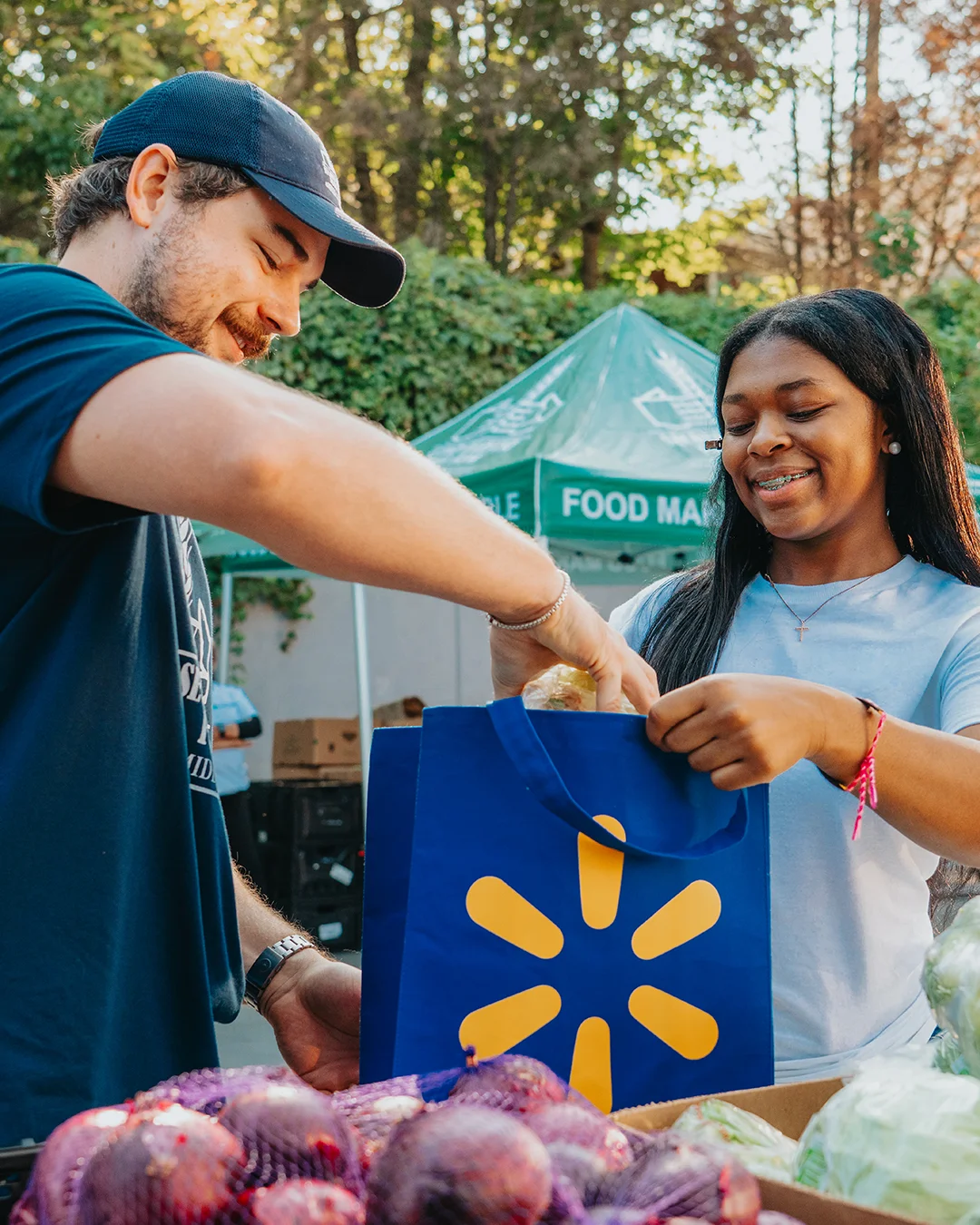Two volunteers are handing out groceries, placing food into a reusable blue shopping bag. They are standing outdoors in front of green tents with text that includes phrases like "FOOD MAKES" and "POSSIBLE." A shopping cart and lush trees are visible in the background, suggesting a community food drive or donation event. The scene feels friendly and collaborative, with bright colors and natural daylight.