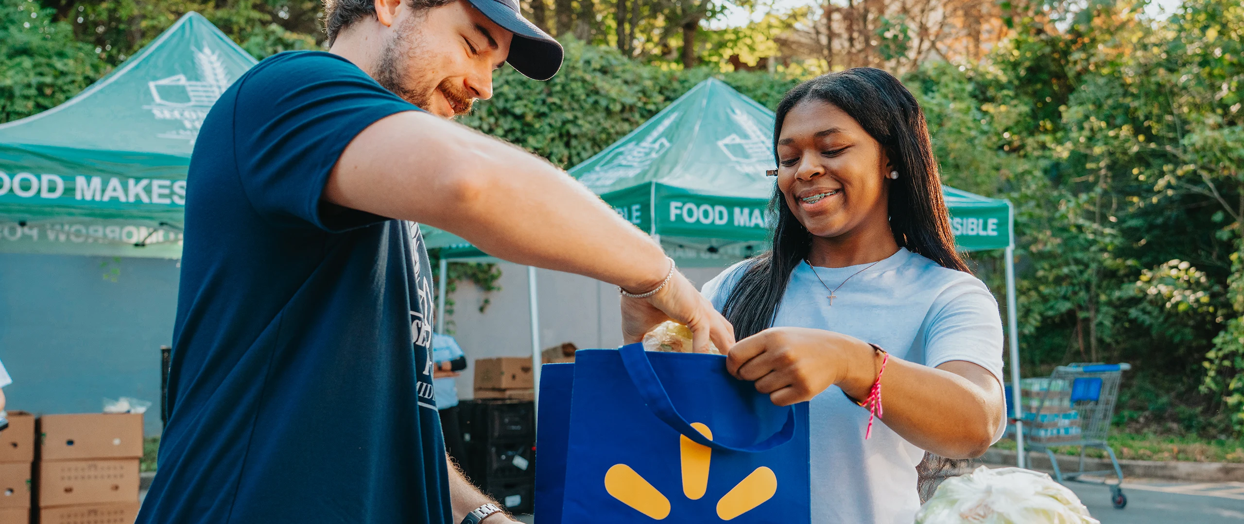 Two volunteers are handing out groceries, placing food into a reusable blue shopping bag. They are standing outdoors in front of green tents with text that includes phrases like "FOOD MAKES" and "POSSIBLE." A shopping cart and lush trees are visible in the background, suggesting a community food drive or donation event. The scene feels friendly and collaborative, with bright colors and natural daylight.