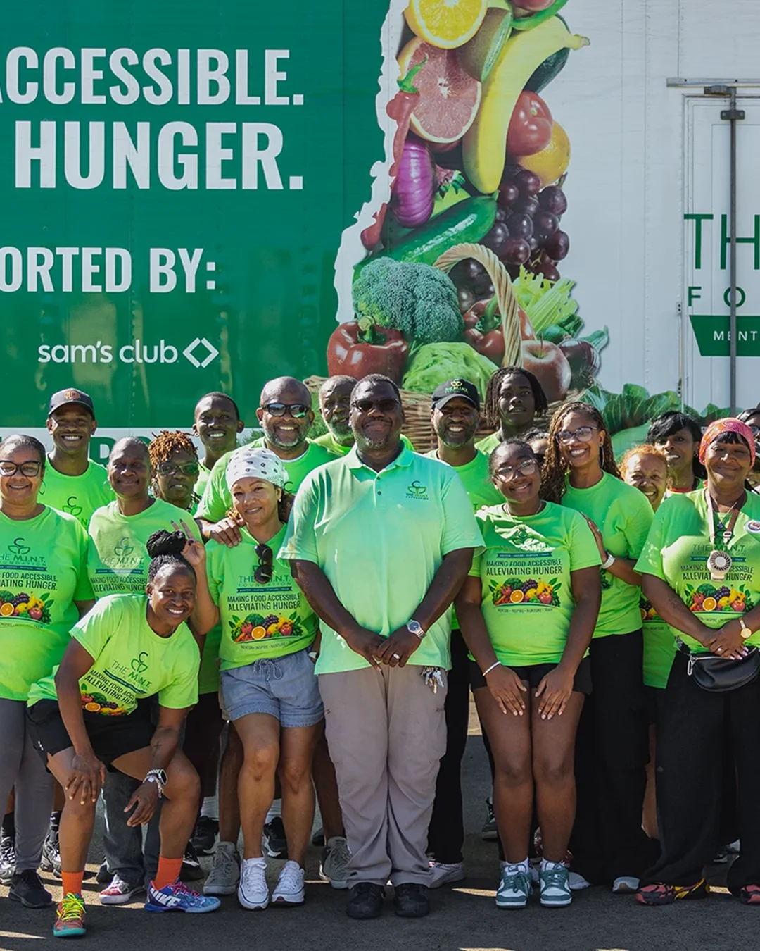 A large group of volunteers in bright green shirts stand together in front of a food distribution truck. The truck side features The M.I.N.T. Foundation name and a message about making food accessible and alleviating hunger. Additional text indicates support from North Texas Food Bank, Walmart, and Sam’s Club. The scene takes place outdoors in a parking area under clear blue skies, conveying community service and charitable outreach.