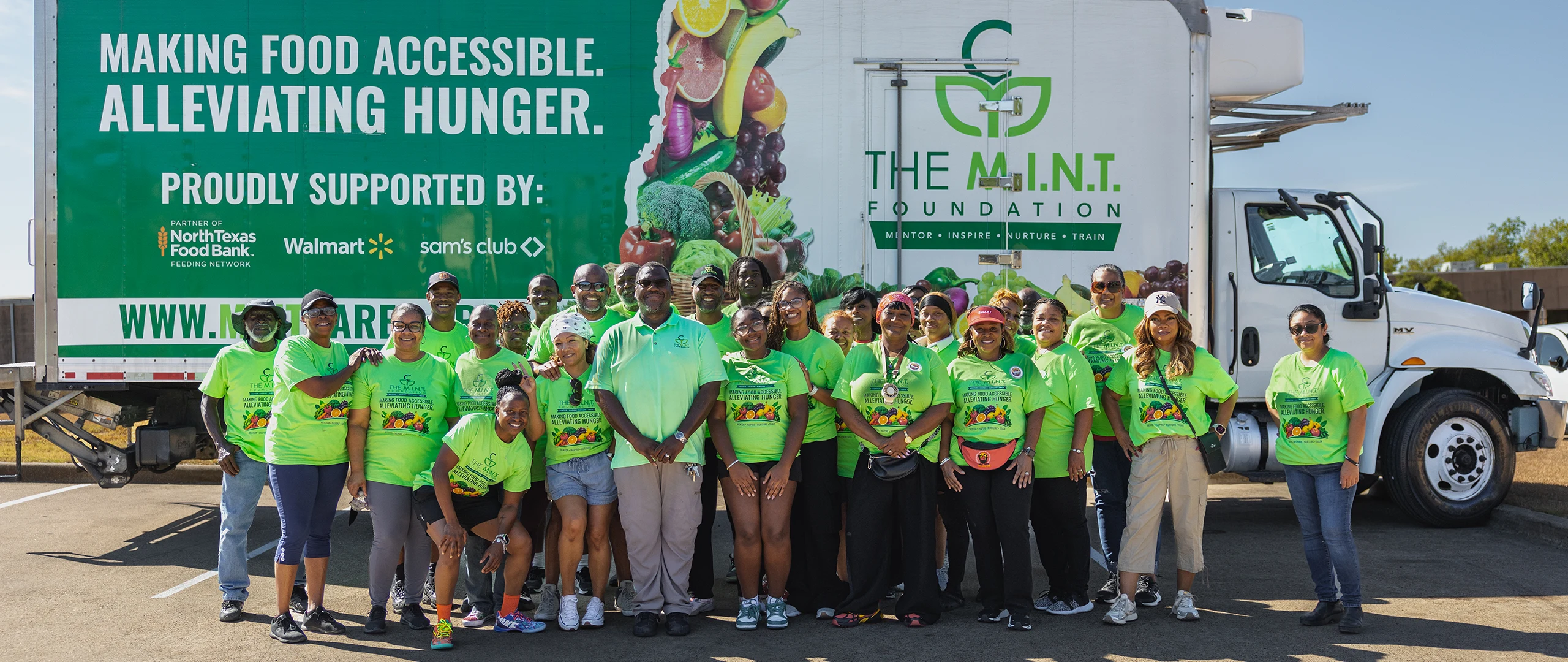 A large group of volunteers in bright green shirts stand together in front of a food distribution truck. The truck side features The M.I.N.T. Foundation name and a message about making food accessible and alleviating hunger. Additional text indicates support from North Texas Food Bank, Walmart, and Sam’s Club. The scene takes place outdoors in a parking area under clear blue skies, conveying community service and charitable outreach.