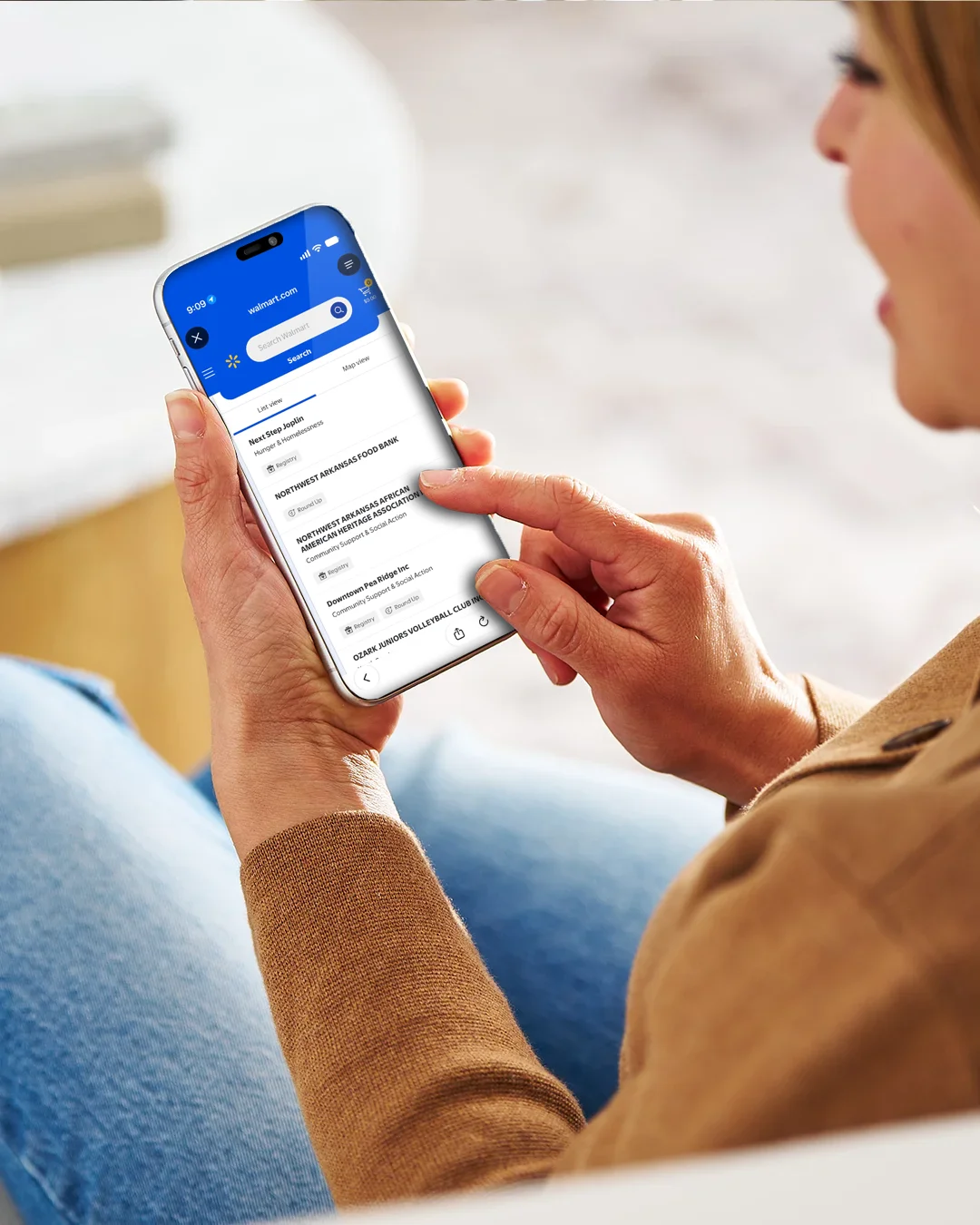 A person holds a smartphone and interacts with a donation or support application on the screen. The interface shows a section titled "Support Organizations on Spark Good" with blue buttons and text fields. The user’s finger is poised to tap a call-to-action button, suggesting online giving or charitable support. The background is softly blurred, keeping focus on the mobile device and the app interface.