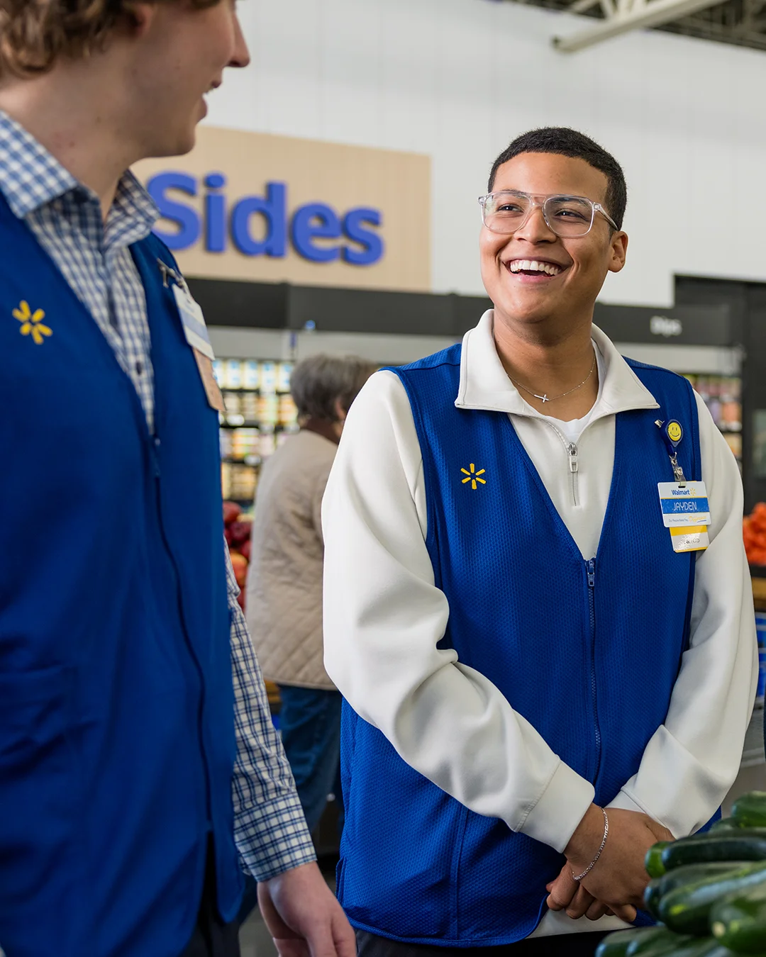 Three store associates wearing blue vests stand together and talk inside a large retail environment. Shelves of products are visible in the background along with overhead signs reading "Sides" and "Pizza." The group appears relaxed and cheerful, suggesting teamwork and a friendly workplace atmosphere. Bright indoor lighting and casual uniforms emphasize an approachable customer service setting.