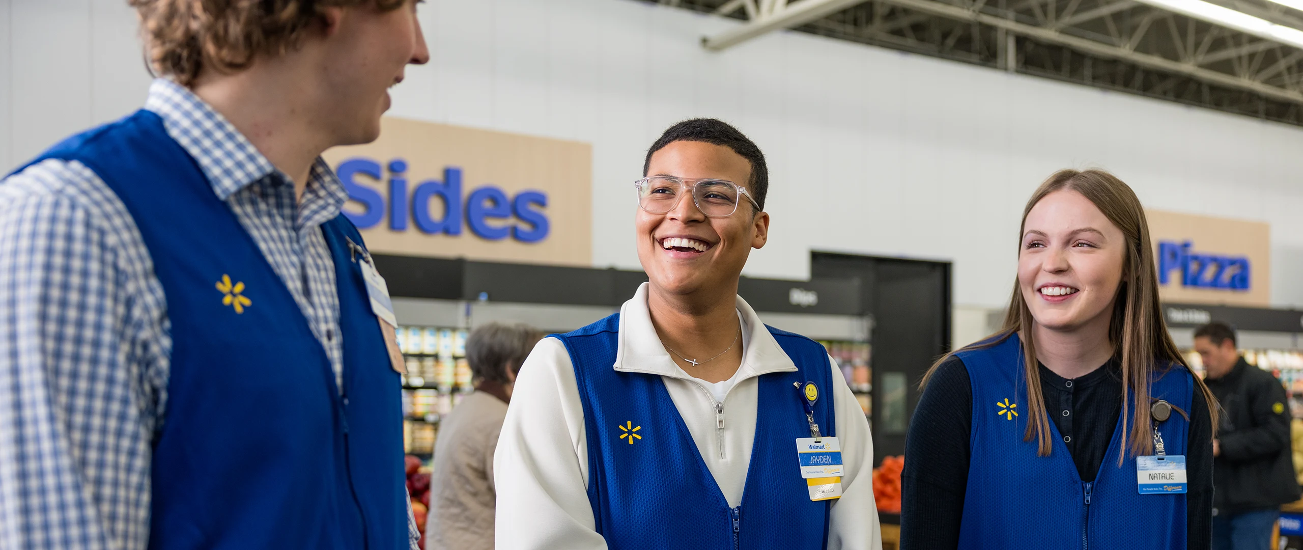 Three store associates wearing blue vests stand together and talk inside a large retail environment. Shelves of products are visible in the background along with overhead signs reading "Sides" and "Pizza." The group appears relaxed and cheerful, suggesting teamwork and a friendly workplace atmosphere. Bright indoor lighting and casual uniforms emphasize an approachable customer service setting.