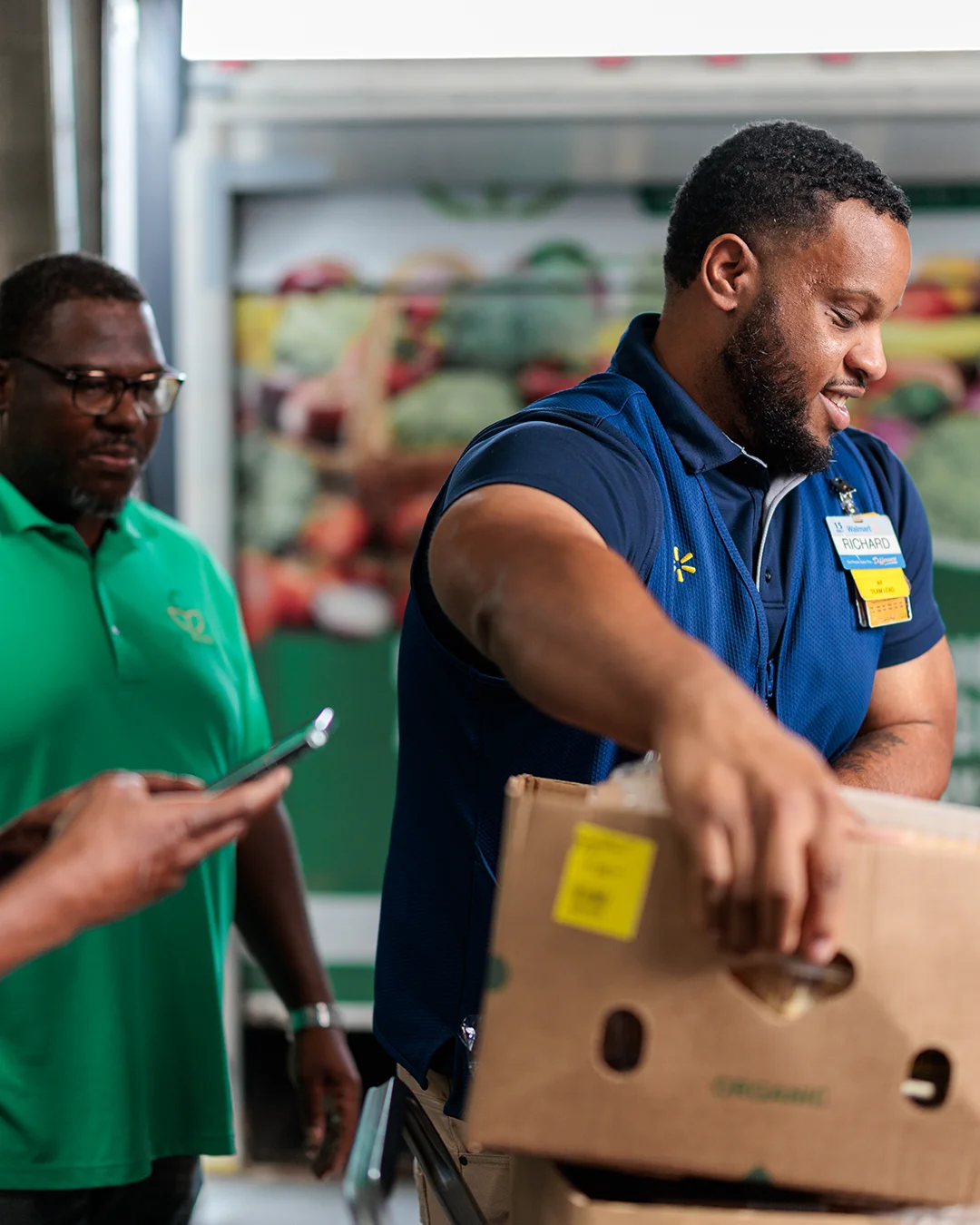 Three workers stand in a grocery or warehouse setting, with one man in the foreground lifting a cardboard box. Another worker checks a smartphone while the third observes, all wearing name badges and work attire. A colorful backdrop of fresh produce is visible behind them, suggesting a food retail environment. The scene conveys teamwork, service, and efficient handling of goods.