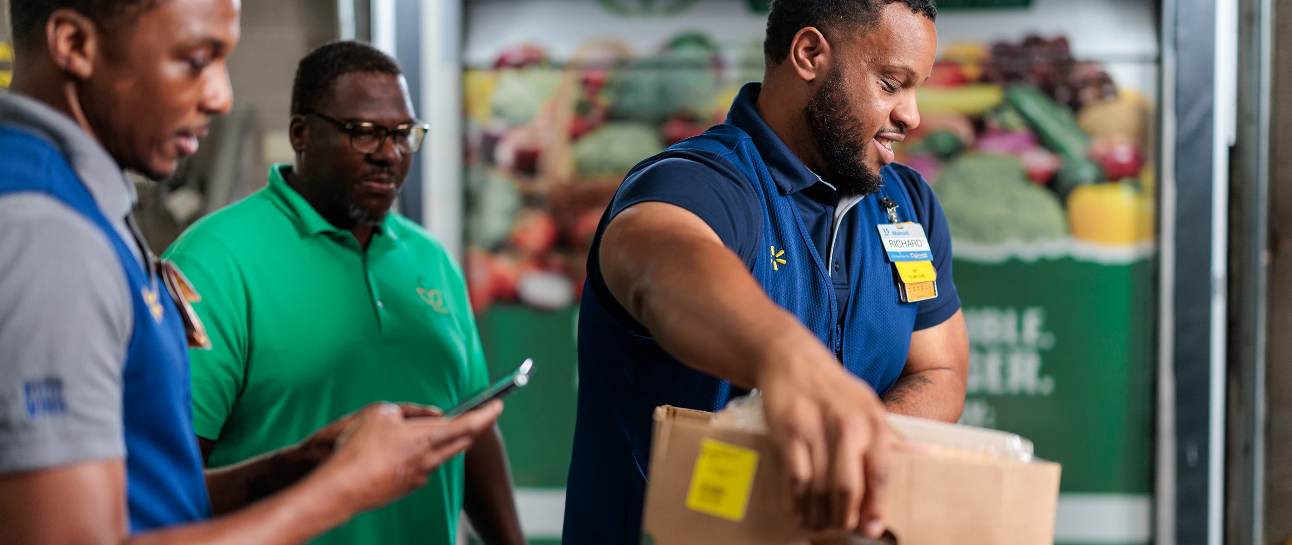 Three workers stand in a grocery or warehouse setting, with one man in the foreground lifting a cardboard box. Another worker checks a smartphone while the third observes, all wearing name badges and work attire. A colorful backdrop of fresh produce is visible behind them, suggesting a food retail environment. The scene conveys teamwork, service, and efficient handling of goods.