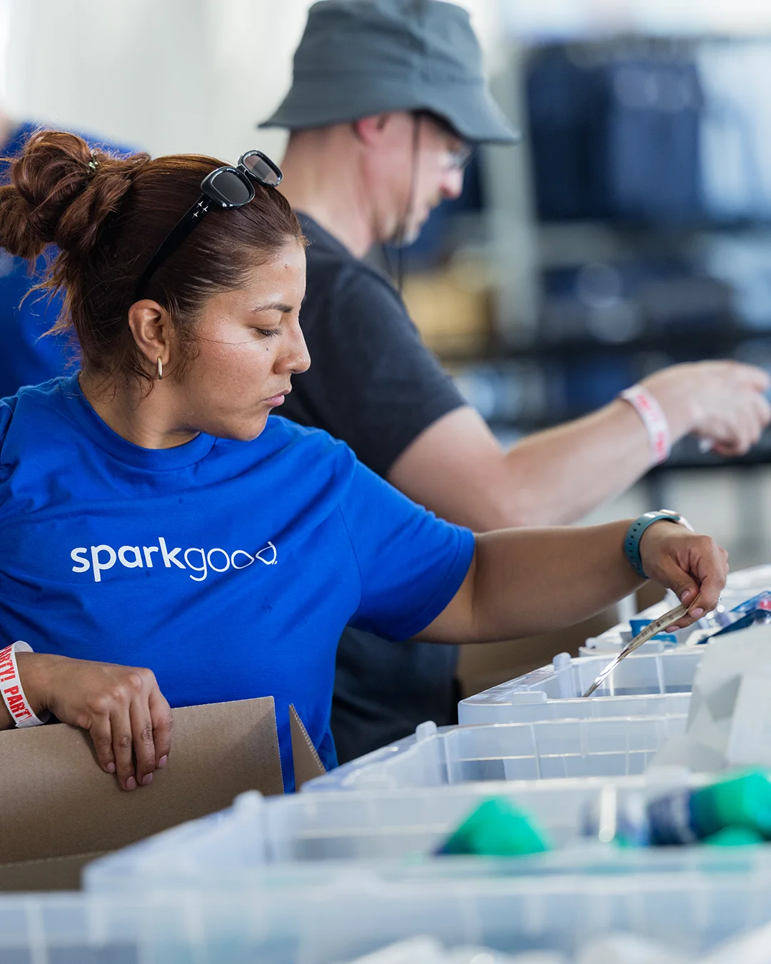 A woman wearing a blue "sparkgood" t-shirt sorts items into plastic bins at an indoor event. Other volunteers work alongside her in the background, creating a busy, collaborative atmosphere. The scene suggests organized community service or charitable work focused on distributing supplies. Bright lighting and casual clothing emphasize an energetic, hands-on environment.