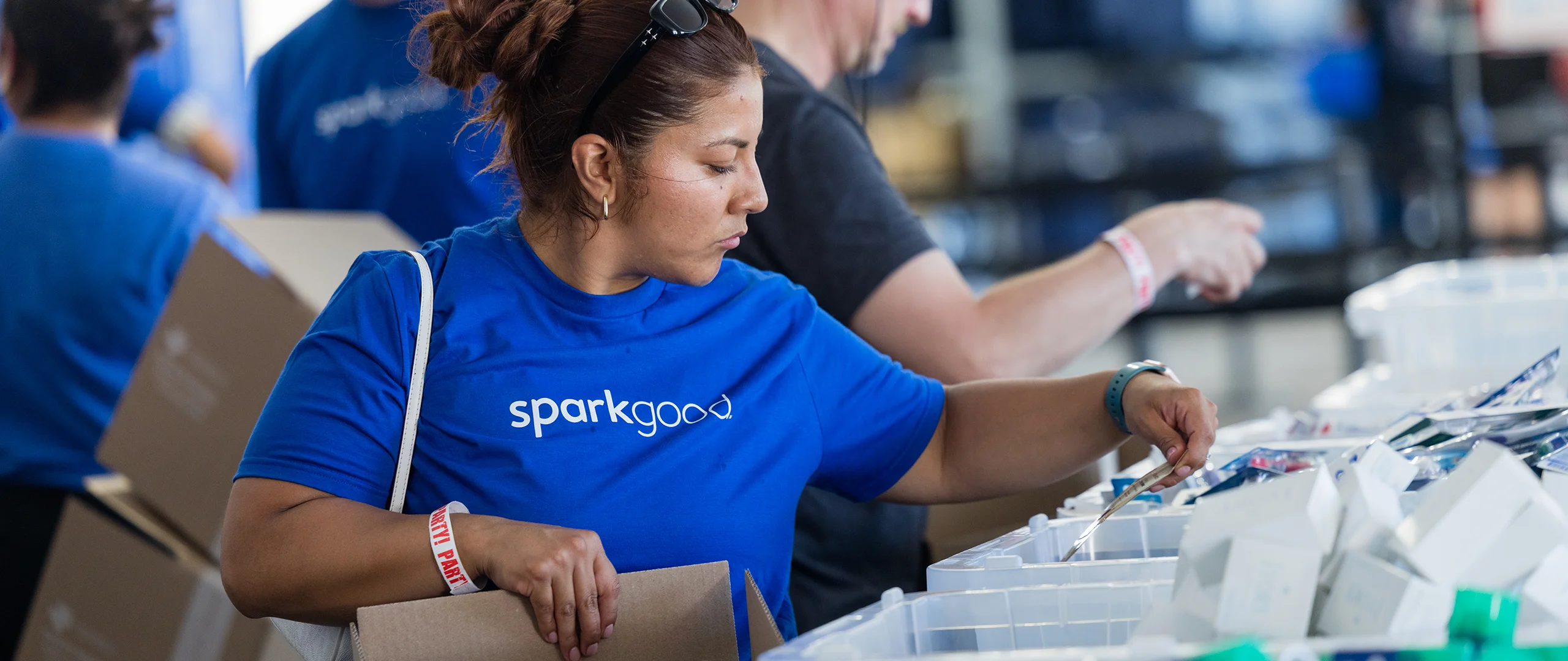 A woman wearing a blue "sparkgood" t-shirt sorts items into plastic bins at an indoor event. Other volunteers work alongside her in the background, creating a busy, collaborative atmosphere. The scene suggests organized community service or charitable work focused on distributing supplies. Bright lighting and casual clothing emphasize an energetic, hands-on environment.