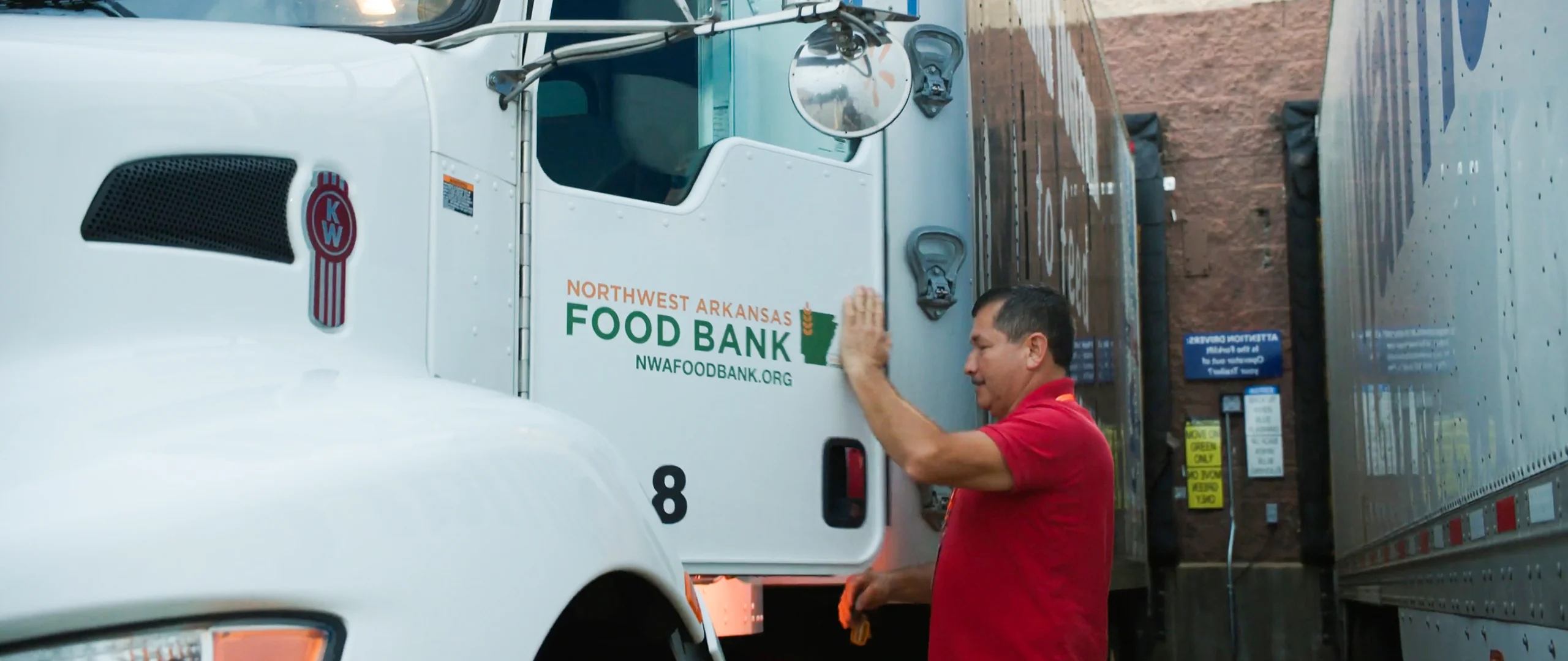 A man in a red shirt is closing the door of a large white truck marked with Northwest Arkansas Food Bank signage. The truck is parked closely beside another trailer in an alley or loading dock area. Text on the truck includes "NORTHWEST ARKANSAS FOOD BANK" and "NWAFoodBank.org," along with the number 8 near the bottom of the door. The scene suggests food distribution or charitable logistics activity.