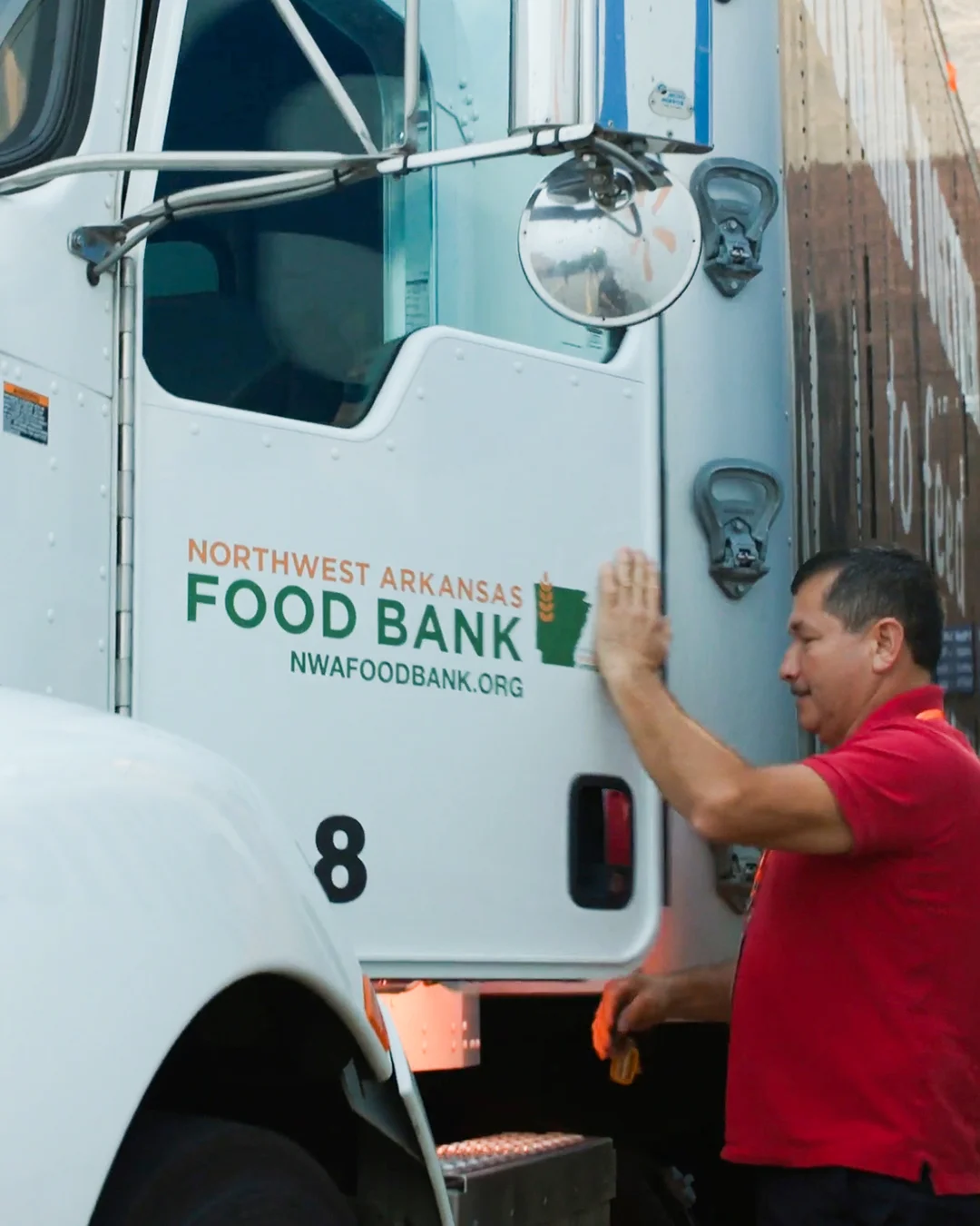 A man in a red shirt is closing the door of a large white truck marked with Northwest Arkansas Food Bank signage. The truck is parked closely beside another trailer in an alley or loading dock area. Text on the truck includes "NORTHWEST ARKANSAS FOOD BANK" and "NWAFoodBank.org," along with the number 8 near the bottom of the door. The scene suggests food distribution or charitable logistics activity.