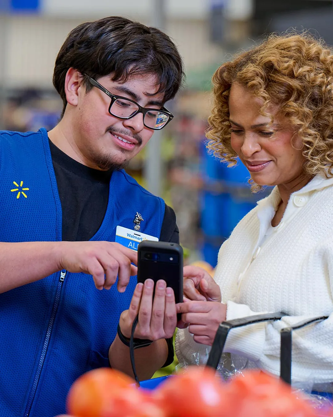 A store employee in a blue vest helps a customer by showing information on a handheld smartphone device. They are standing together in a grocery aisle with fresh produce visible in the background. The interaction appears friendly and supportive, suggesting customer service and in-store assistance. The environment is bright and organized, emphasizing a modern retail shopping experience.