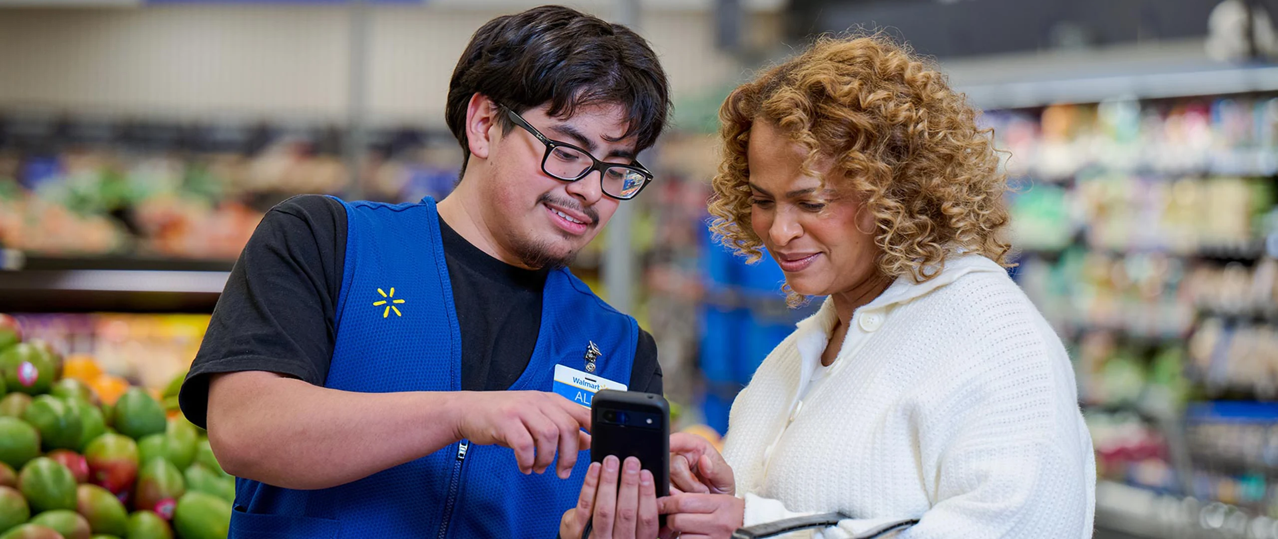 A store employee in a blue vest helps a customer by showing information on a handheld smartphone device. They are standing together in a grocery aisle with fresh produce visible in the background. The interaction appears friendly and supportive, suggesting customer service and in-store assistance. The environment is bright and organized, emphasizing a modern retail shopping experience.