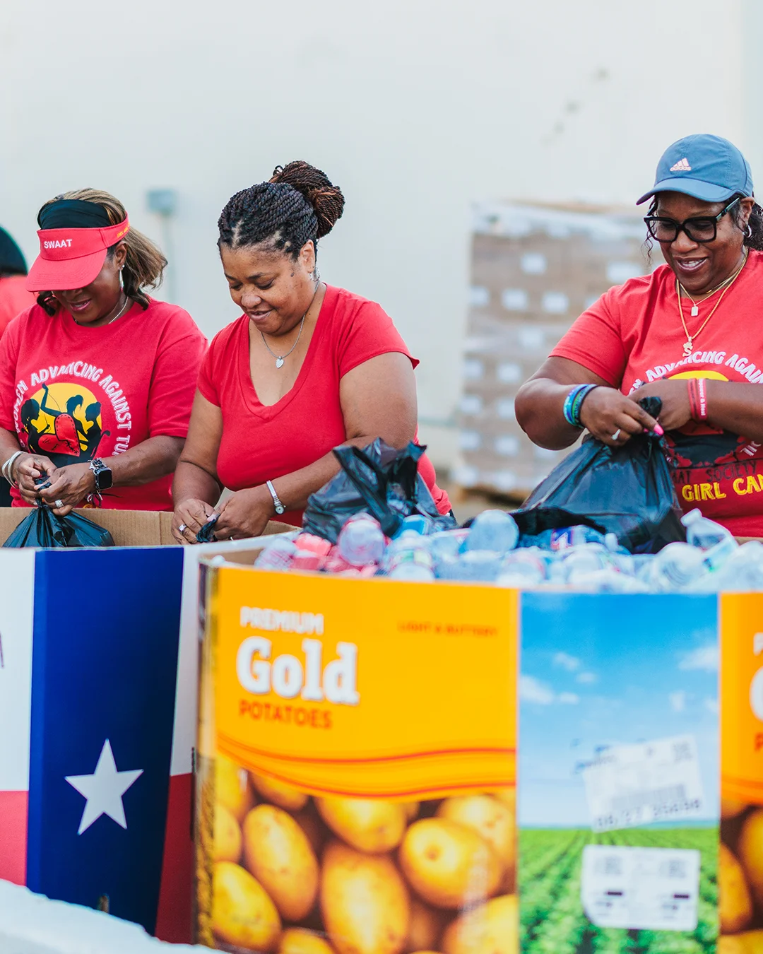Three women in red shirts work together to pack food items into black bags at an outdoor community event. They stand behind large cardboard bins labeled with visible text including "Lone Star Potatoes" and "Premium Gold Potatoes." The setting appears to be a warehouse or distribution area with stacked boxes and supplies in the background. The scene conveys teamwork, service, and charitable support for the local community.