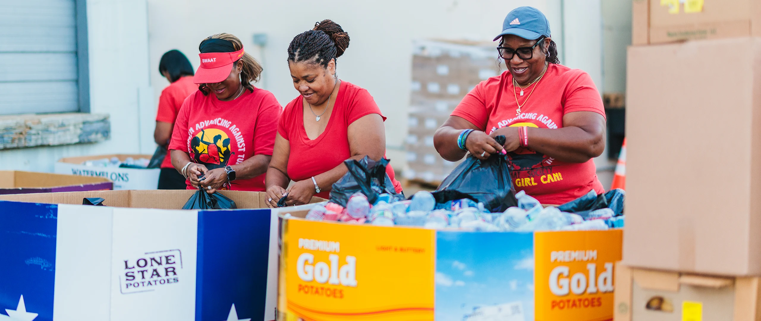 Three women in red shirts work together to pack food items into black bags at an outdoor community event. They stand behind large cardboard bins labeled with visible text including "Lone Star Potatoes" and "Premium Gold Potatoes." The setting appears to be a warehouse or distribution area with stacked boxes and supplies in the background. The scene conveys teamwork, service, and charitable support for the local community.