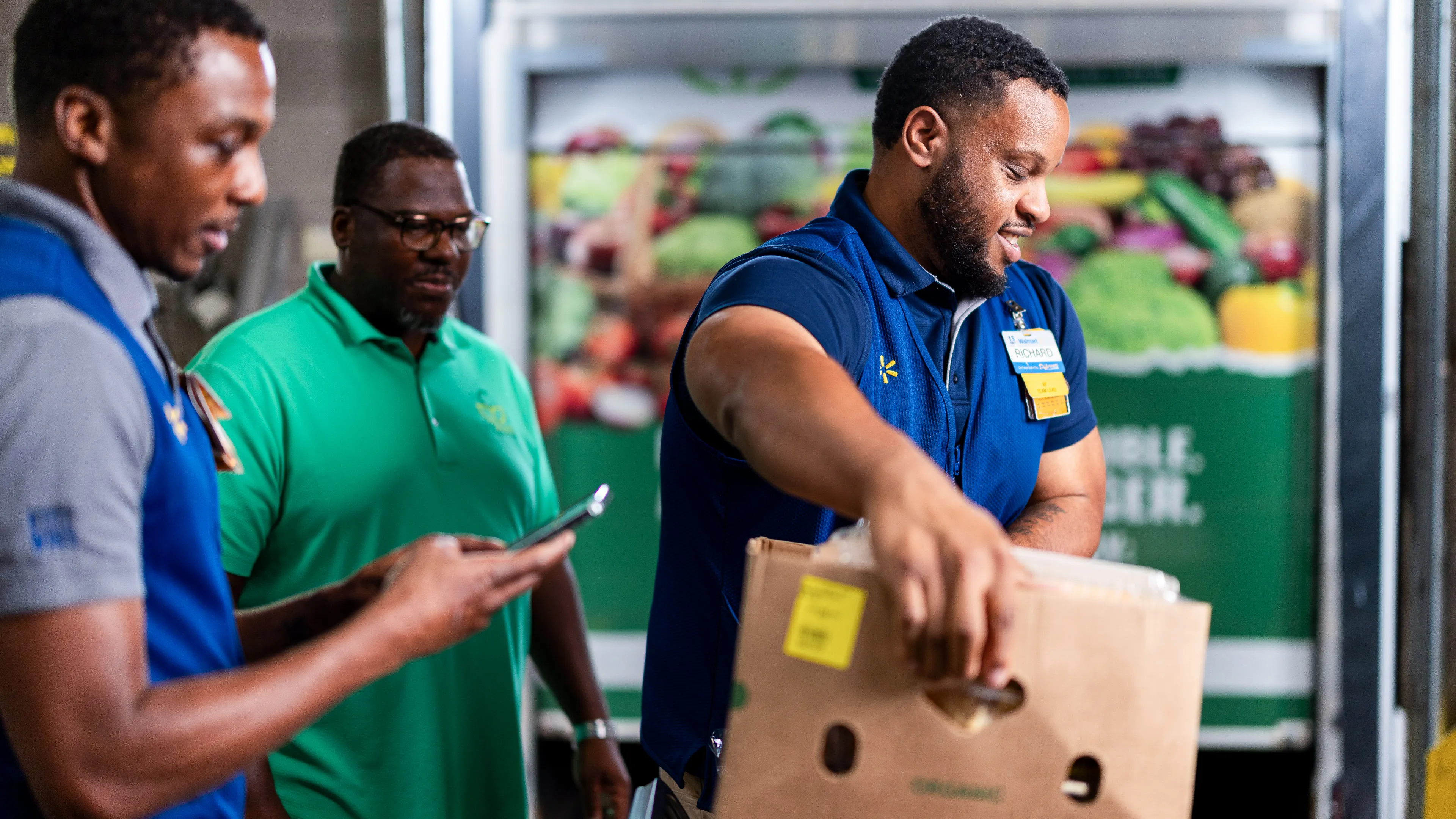 Three male workers are shown in a grocery or warehouse setting, with one man lifting a large cardboard box labeled "ORGANIC." Another worker checks a smartphone while a third stands nearby, and a refrigerated display of colorful fruits and vegetables is visible in the background. The men wear polo shirts and name badges, suggesting a professional retail or fulfillment environment. The scene conveys teamwork, productivity, and fresh food handling.