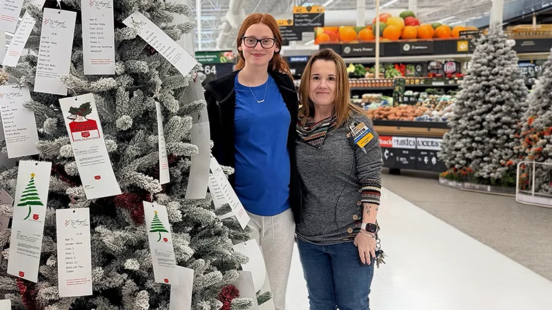 Two women stand together next to a flocked Christmas tree covered with hanging wish list cards. The scene appears inside a large grocery or retail store, with produce and other merchandise visible in the background. One woman wears a blue shirt and light pants, while the other wears a gray top with a name badge and lanyard. The decorated tree and additional trees in the distance create a festive holiday atmosphere.