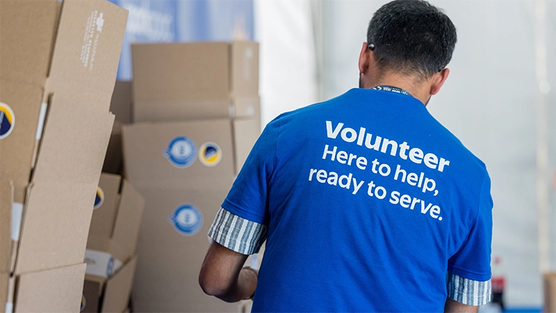 A man wearing a blue T-shirt with the word "Volunteer" printed on the back stands among stacked cardboard boxes. The shirt text reads "Volunteer Here to help, ready to serve," emphasizing community support and service. The setting appears to be an indoor event or distribution center with multiple brown boxes in the background. The overall scene suggests organized volunteer work and charitable assistance.