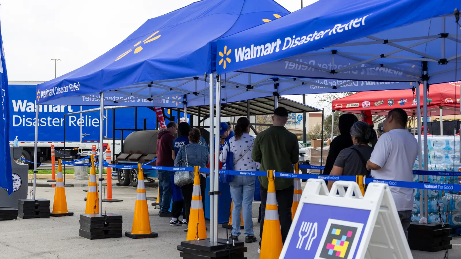 People stand in line under blue Walmart Disaster Relief tents at an outdoor assistance site. Safety cones and barrier tape organize the queue area, while staff work at tables inside the canopies. A large Walmart Disaster Relief truck is visible in the background, reinforcing the emergency support setting. Nearby signage with food-related icons suggests distribution of supplies or meals to affected residents.