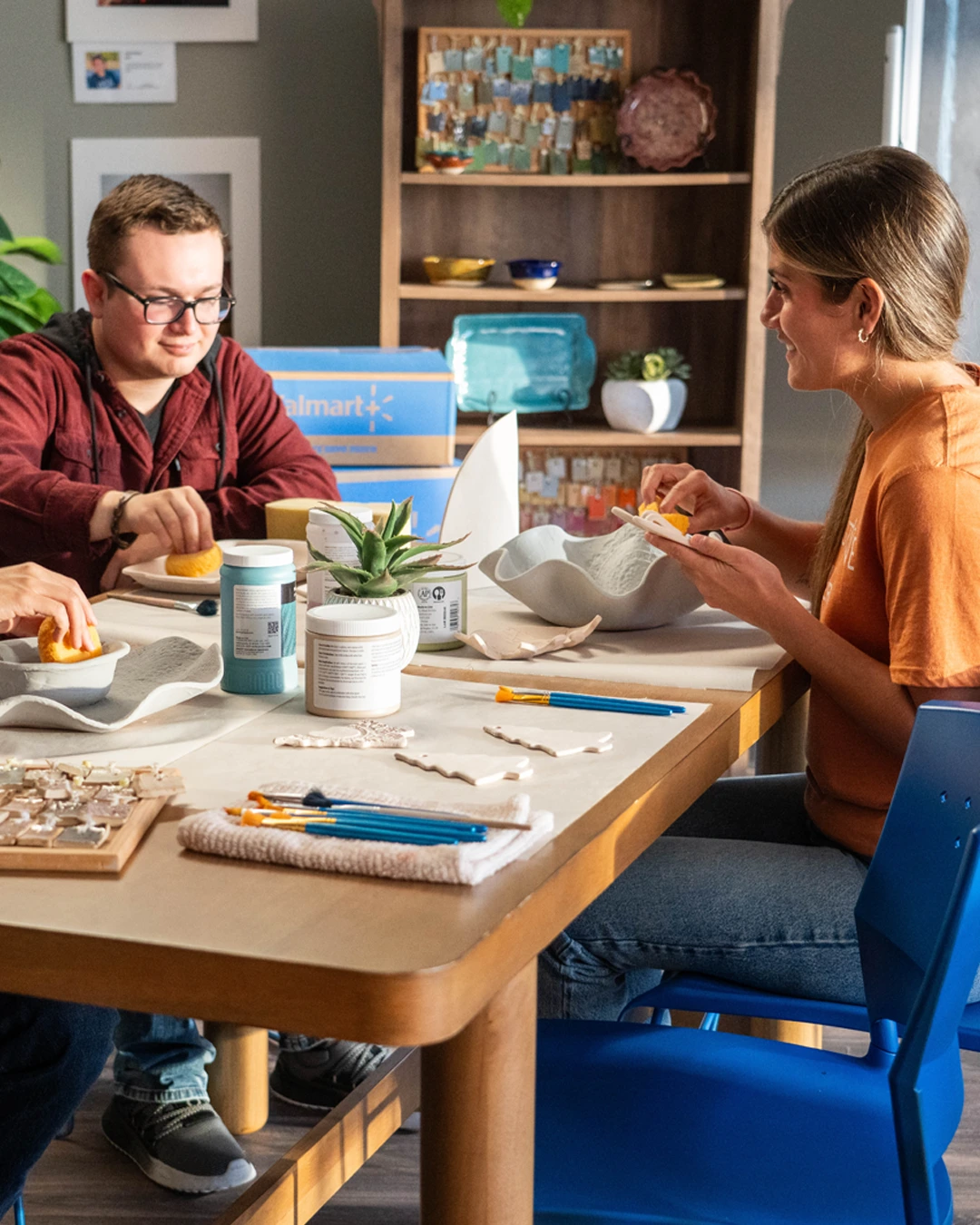 Three young adults sit around a wooden table engaged in a creative craft activity. They are arranging small tiles and materials into designs, with brushes, containers, and tools spread across the tabletop. The setting appears to be a bright, modern room with plants, shelves, and a chalkboard in the background. Warm sunlight and casual clothing create a relaxed, friendly atmosphere focused on art and collaboration.