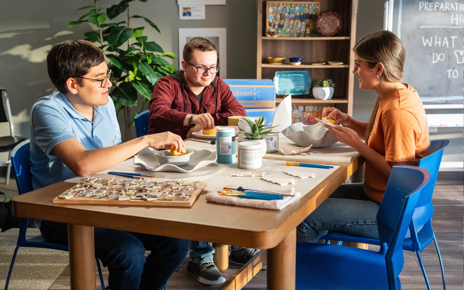 Three young adults sit around a wooden table engaged in a creative craft activity. They are arranging small tiles and materials into designs, with brushes, containers, and tools spread across the tabletop. The setting appears to be a bright, modern room with plants, shelves, and a chalkboard in the background. Warm sunlight and casual clothing create a relaxed, friendly atmosphere focused on art and collaboration.