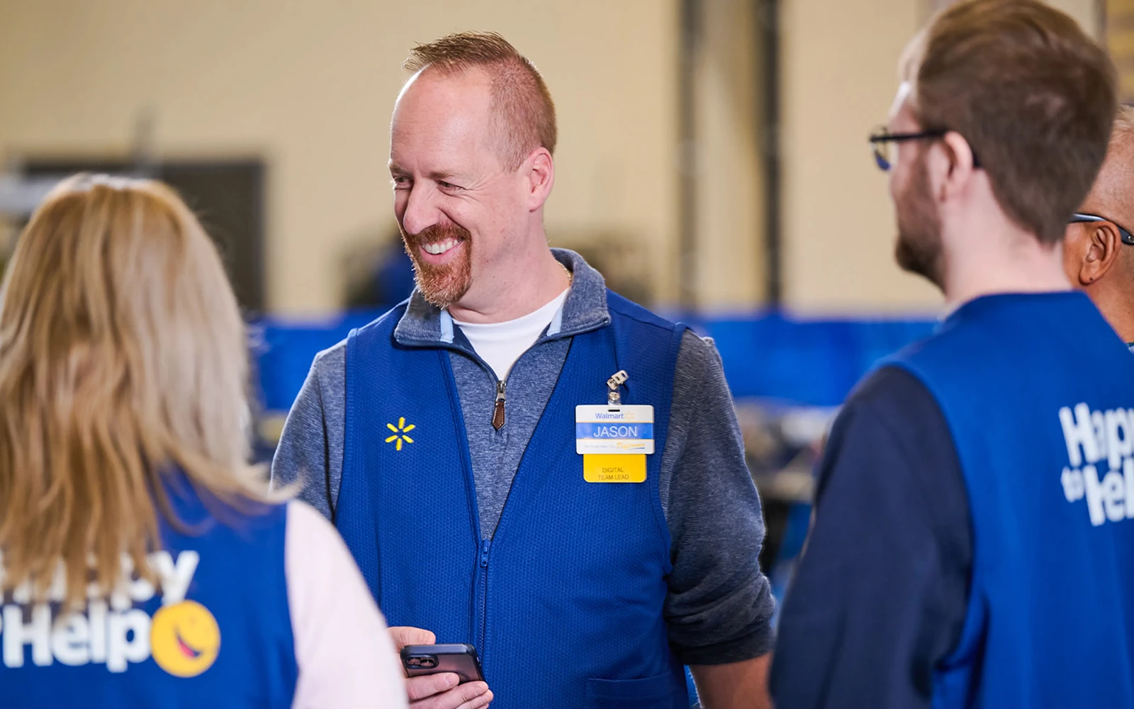 A group of retail store associates wearing blue vests stand together and talk. The central man smiles while holding a smartphone, engaging with coworkers in a brightly lit store or warehouse setting. Name badges and printed text are visible on the vests, indicating a customer service role. The background shows an indoor commercial environment with shelves and equipment slightly out of focus.