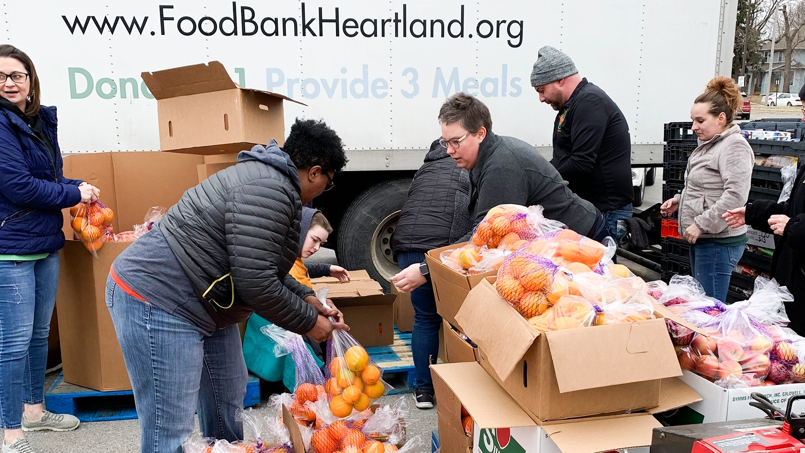 A group of volunteers sort and distribute bags of oranges beside a large truck. The truck features the visible website text "FoodBankHeartland.org" and a donation message in the background. Cardboard boxes and crates are filled with fresh produce on a pallet in an outdoor setting. People in casual winter clothing work together, creating a busy and collaborative atmosphere.