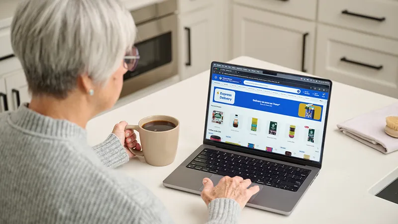 An older woman with short gray hair sits at a kitchen counter using a laptop computer. She is browsing an online grocery delivery website that displays various food products in a grid layout. The woman holds a mug of coffee in one hand while navigating the site with the other. Bright kitchen cabinets and countertops create a clean, modern home setting.