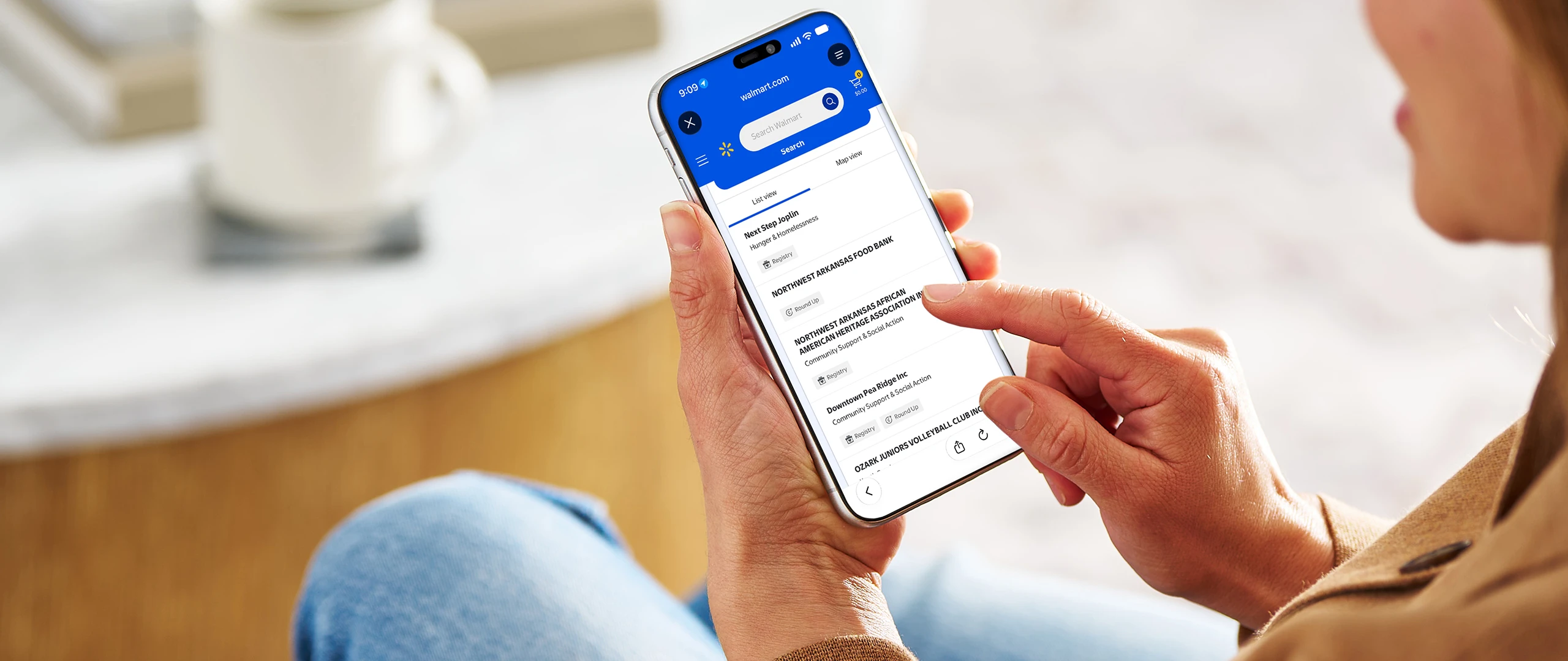 A woman holds a modern smartphone while seated indoors, interacting with a blue-and-white mobile app interface. She appears to be reviewing financial or account information on the screen in a relaxed home setting. The background is softly blurred, highlighting the device and her hand. Warm lighting and casual clothing contribute to a comfortable, everyday lifestyle atmosphere.