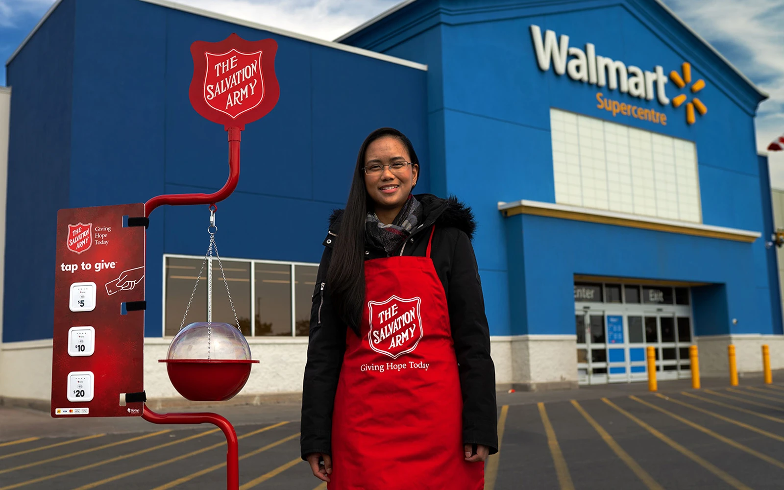 A Salvation Army volunteer wearing a red apron stands in front of a Walmart Supercentre entrance. Beside her is a red donation stand with a hanging collection kettle and a tap-to-give panel. The scene takes place in a parking lot with yellow bollards and the blue Walmart storefront in the background. Text on the apron reads "Giving Hope Today," and the Salvation Army shield is clearly visible on the stand and apron.