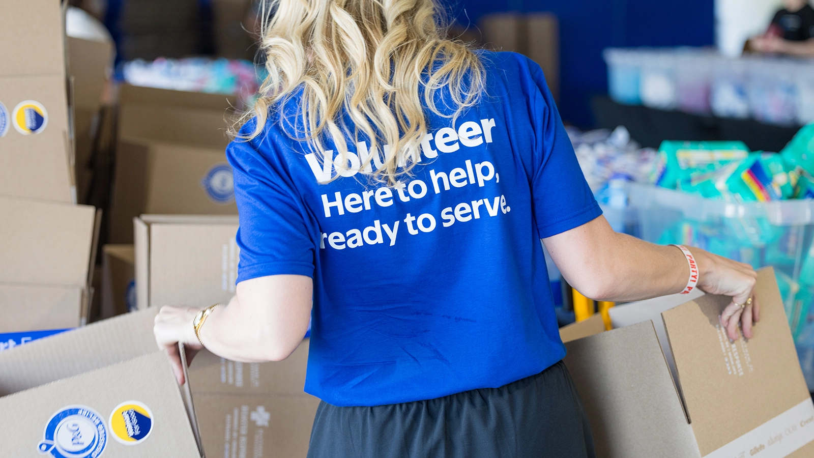 A volunteer wearing a bright blue T-shirt is lifting and arranging cardboard boxes in a busy indoor setting. The back of the shirt reads "Volunteer Here to help, ready to serve." Numerous boxes and supplies are visible around the person, suggesting a community service or donation drive. The scene conveys teamwork, generosity, and active participation in helping others.