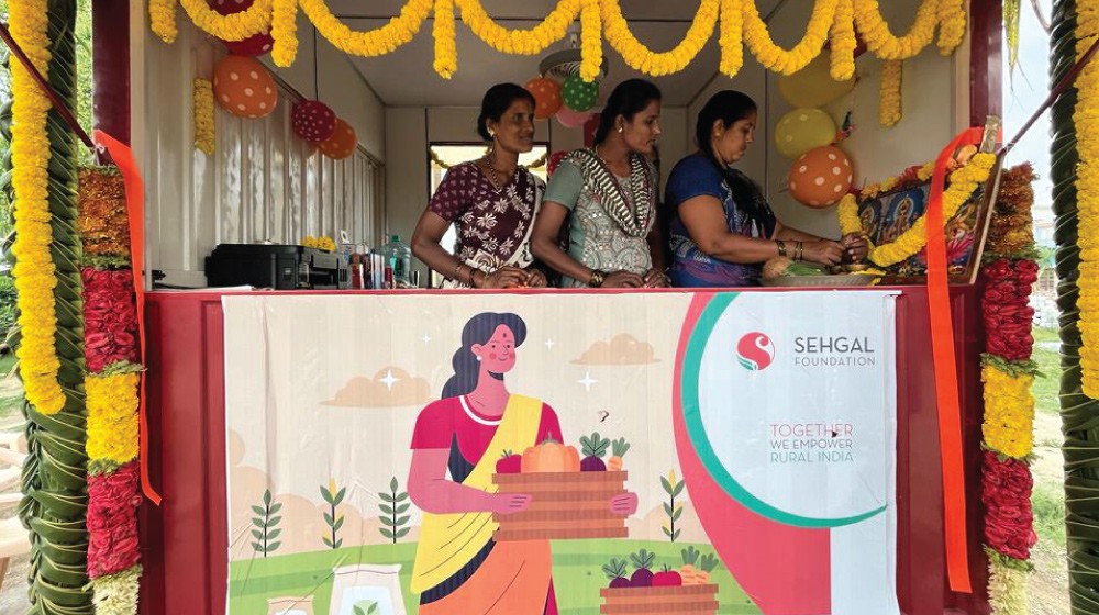 Three women stand in a Sehgal Foundation booth. The booth text says "Sehgal Foundation. Together we empower rural India."
